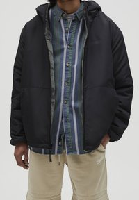 Czarna pikowana kurtka z kapturem i zamkiem błyskawicznym z przodu, noszona na niebieskiej i zielonej koszuli w paski z guzikami. Posiada miękką podszewkę i elastyczne mankiety.