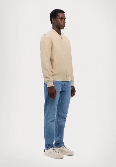 Giacca bomber beige con zip frontale e polsini a coste, abbinata a jeans azzurri e sneakers crema con dettagli neri.