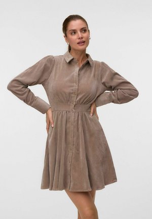Femme portant une robe marron à manches longues boutonnée avec une taille cintrée, debout les mains sur les hanches et regardant sur le côté.