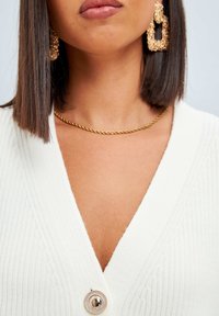 Vit stickad cardigan med djup V-ringning, med en stor rund knappstängning. Guldvridet halsband och glittrande hängande örhängen.