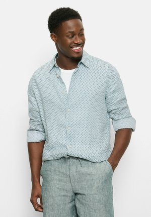 Man in een lichtblauw overhemd met patroon en opgerolde mouwen, en lichtblauwe linnen broek, glimlachend en naar beneden kijkend met hand in de zak.