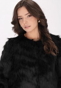 Zwarte faux fur jas met een pluizige textuur en ronde halslijn, gecombineerd met een gouden ketting. Model heeft golvend bruin haar.