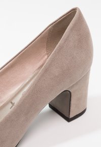Chaussure à talon haut en daim beige avec un talon bloc et un bout arrondi, présentant une texture lisse et un léger éclat sur la doublure intérieure.