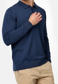 Hombre ajustando el cuello de una camisa polo de manga larga de color azul marino, vestido con pantalones beige, de pie contra un fondo blanco.