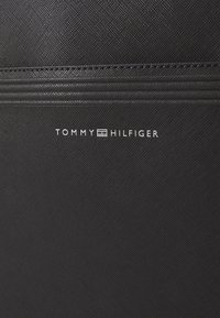 Fekete textúrázott anyag, amelyen fehér Tommy Hilfiger logo található, a lenti részen elhelyezve, felette vízszintes varrásokkal.