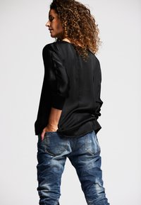 Schwarzes, glattes Oberteil mit langen Ärmeln, kombiniert mit verwaschenen, strukturierten blauen Jeans. Das Modell wird von hinten gezeigt, mit Händen in den Taschen.