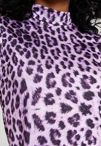 Suéter de cuello alto morado con estampado de leopardo negro, usado por una persona con cabello rizado oscuro visible en el cuello.