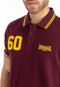 Burgundy pólóing sárga kiegészítőkkel, hímzett "60" és "Lonsdale London" logókkal, gombos lezárással és bordás gallérral.