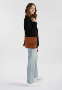 Borsa a spalla in pelle marrone con chiusura a pattina, design piatto e tracolla regolabile, indossata in modo informale con jeans azzurri chiari e sneakers bianche.