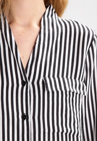 Camisa a rayas verticales en blanco y negro con cuello de solapa entallado, un bolsillo en el pecho y botones negros sobre una tela lisa.