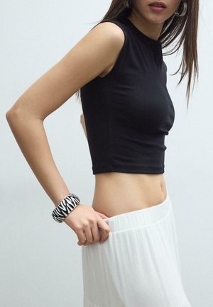 Donna che indossa un top corto nero senza maniche e una gonna bianca, mentre aggiusta la vita, con un braccialetto a motivo bianco e nero al polso.