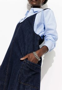 Salopette en denim à coupe large, de couleur bleu foncé, avec une poche avant. Associée à une chemise à rayures bleues claires à boutons. Bracelet en argent au poignet.