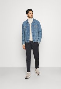 Chaqueta de mezclilla en azul claro, cuello alto en crema, pantalones oscuros y zapatillas beige. Presenta un diseño clásico con dos bolsillos frontales.