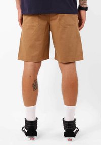 Shorts en coton beige avec une coupe décontractée, comportant deux poches arrière. Portés avec des baskets montantes noires et des chaussettes blanches.