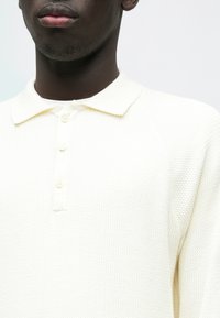 Polo branca feita de tecido texturizado, com decote com gola e três botões na frente. Design simples e limpo.