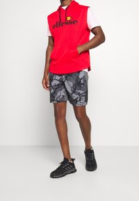 Red sleeveless hoodie with black logo, paired with gray camouflage shorts. Black athletic shoes with textured finish and laces.