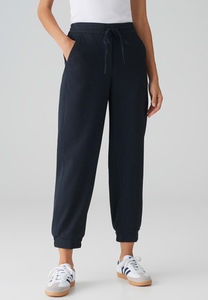 Pantalons de survêtement bleu marine avec une taille à cordon, comportant des poches latérales et des poignets élastiques. Le tissu a une finition texturée, associé à des baskets blanches.
