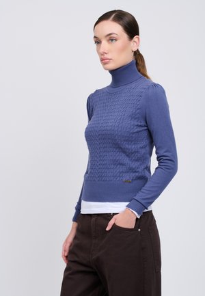 Femme aux cheveux bruns attachés en queue de cheval, portant un pull col roulé bleu torsadé et un pantalon foncé, debout avec une main dans la poche.