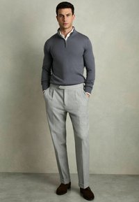 Grau zip-up Pullover, kombiniert mit hellgrauen, plissierten Hosen. Das Model steht mit den Händen in den Taschen und trägt dunkelbraune Schuhe. Glatte Texturen überall.