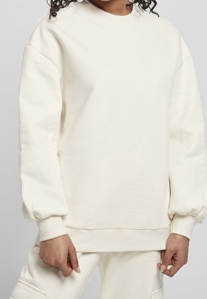 Personne portant un sweat crème surdimensionné et un pantalon assorti, debout, les mains détendues le long du corps, devant un fond uni.