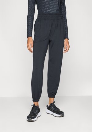 Person wearing black elastic-waist jogger pants with cuffed ankles and black and white sports shoes on a plain light background.