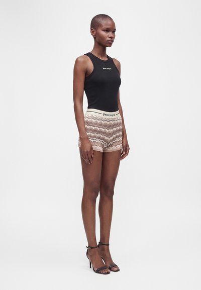 Femme aux cheveux courts portant un haut noir sans manches et un short beige à motifs, debout dans des sandales noires à talons sur un fond uni.