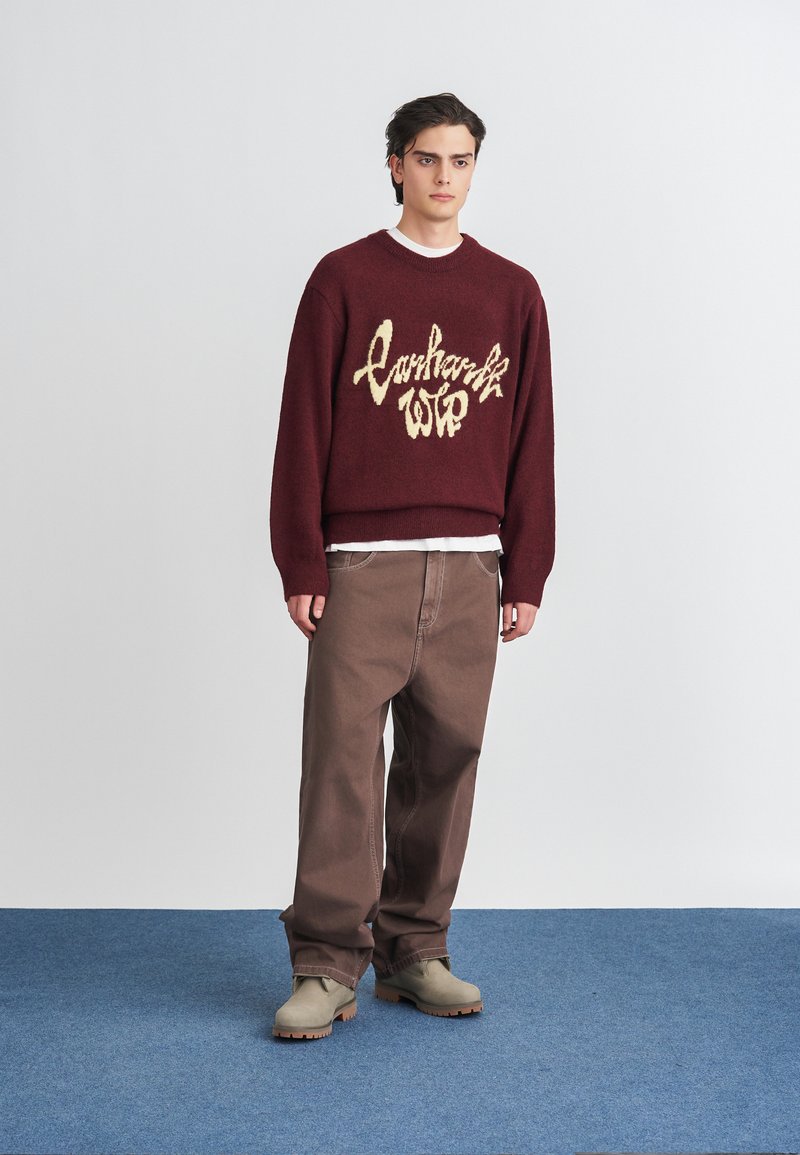 Burgundi kötött pulóver krémszínű logóval, laza barna nadrággal és bézs klumpákkal párosítva, kék szőnyegen állva, fehér háttér előtt.