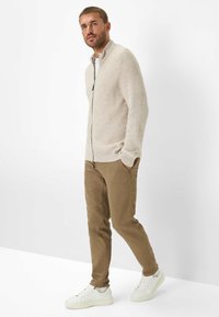 Beige gestreifter Strick-Zipfelpullover, beige Chinos und weiße Sneakers. Das Modell hat die Hände in den Taschen und steht vor einem weißen Hintergrund.