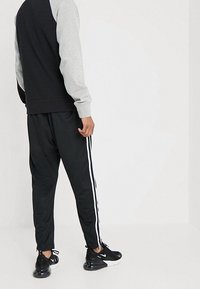 Person som bär svarta träningsbyxor med vita sidostäck, svartvita sneakers och en svart och grå långärmad topp, som står mot en enfärgad bakgrund.