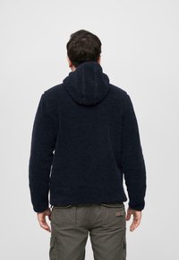 Haut à capuche en polaire bleu marine avec une surface texturée et douce, doté d'un cordon de serrage. Présente une coupe décontractée et des poignets élastiques.