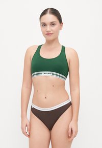Žalios sportinės liemenėlės su baltos ir tamsiai žalios juostos akcentu, su 'LACOSTE' logotipu. Rudos bikinio apatinės dalies, su baltu logotipu ant juosmens.