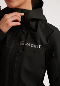 Giacca nera impermeabile con colletto alto, cerniere e il nome del marchio "JACKIT" in argento sul davanti. Texture liscia.