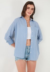 Blue and white striped button-up shirt with a collar, long sleeves, and a relaxed fit, paired with frayed denim shorts.