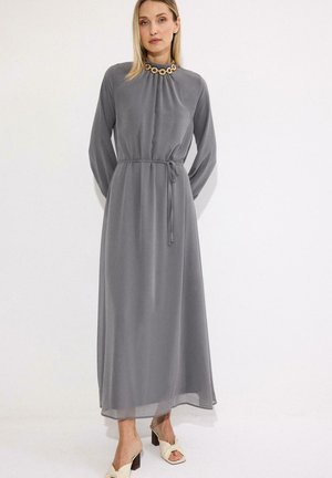 Γυναίκα που φοράει ένα μακρύ γκρι φόρεμα με σφιγμένη μέση και ένα χρυσό αλυσίδα κολιέ, σε συνδυασμό με μπεζ σανδάλια με τακούνι, στέκεται μπροστά από έναν λευκό τοίχο.