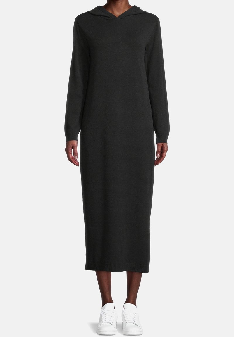 Robe midi noire à manches longues avec capuche portée avec des baskets blanches sur une personne debout sur un fond blanc uni.