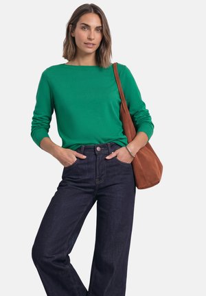 Femme portant un haut vert à manches longues et un jean foncé, les mains dans les poches, portant un sac à bandoulière marron sur un fond uni.