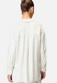 White long-sleeve shirt with vertical stripes, featuring a buttoned cuff, relaxed fit, and soft texture. Back pleat for added movement.
