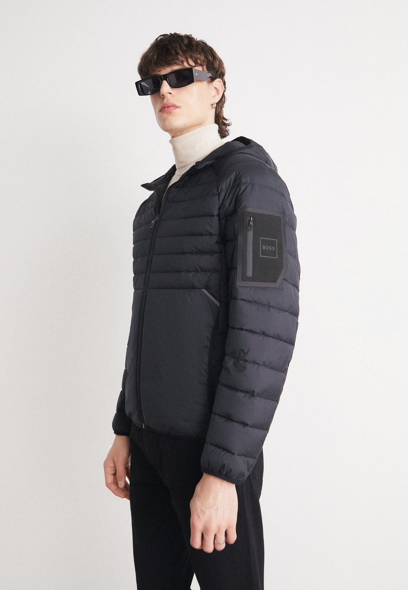 Svart pufferjacka med horisontell kviltad design, huva och dragkedjefickor på ärmarna med logotypapplikation. Materialet verkar slätt och glänsande.