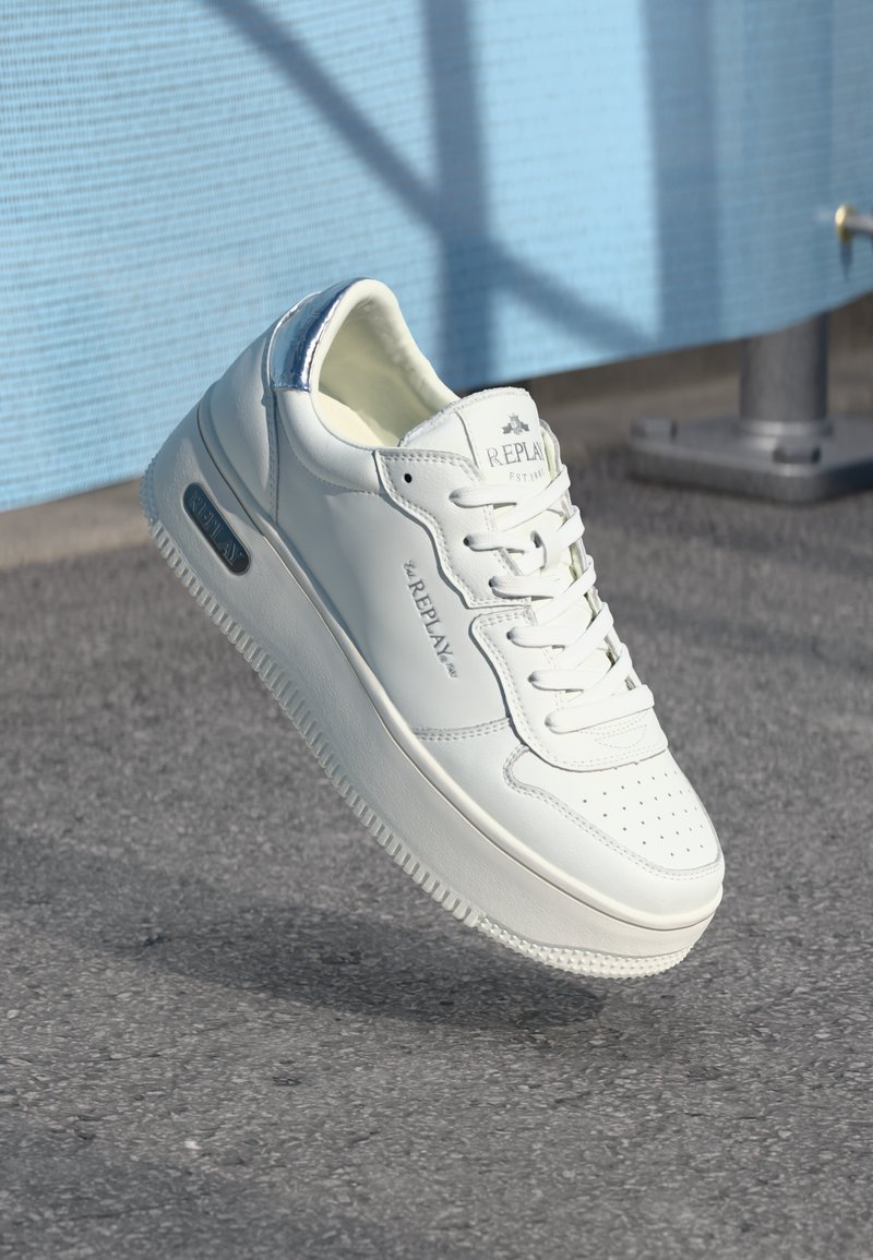Sneaker Replay blanc avec lacets et détail de talon métallique bleu, flottant au-dessus de l'asphalte avec un fond de mur texturé bleu.