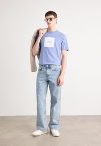 T-shirt de algodão lavanda com logótipo quadrado branco, combinada com calças de ganga azul claro e sapatilhas brancas. Modelo segurando uma jaqueta bege.