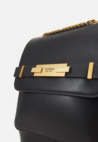Černá kožená kabelka s kovovým zlatým zapínáním s nápisem "GUESS EST 1981" a řetízkovým popruhem pro nošení. Hladká textura, strukturovaný tvar.