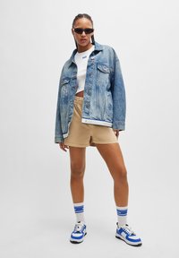 Licht denim oversized jack, witte cropped top, khaki shorts en blauw/witte sneakers. Accessoires zijn slanke zonnebril en gestreepte sokken.