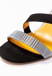 Svart mockaskandalsandal med två korsremmar; en är i svart tyg och den andra har ett rutmönster i grått och blått. Gul innersula.