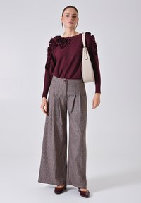 Burgunderfarbener Pullover mit langen Ärmeln und Rüschendetails; breite karierte Hose in Brauntönen; beigefarbene Handtasche; weinrote Schuhe.