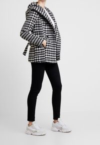 Cappotto in bianco e nero con motivo a denti di cane, con colletto ampio, due tasche laterali e vita cintata, abbinato a pantaloni neri e sneakers bianche.