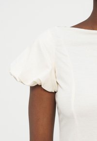 Δεξί χέρι ατόμου με σκουρόχρωμο δέρμα, που φορά άσπρο τοπ με κοντά, φουσκωτά μανίκια πάνω σε ένα απλό ανοιχτό φόντο.