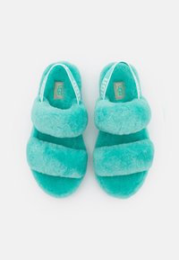 Tyrkysové chlupaté pantofle se dvěma širokými pásky a měkkou texturou. Na popruzích je značka UGG. Lehké, pohodlné obuvnické provedení.
