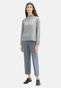 Pulover gri tricotat cu guler înalt, cu manșete ondulate, asortat cu pantaloni bleumarin croiți și pantofi negri cu șireturi; modelul stă în fața unui fundal alb.