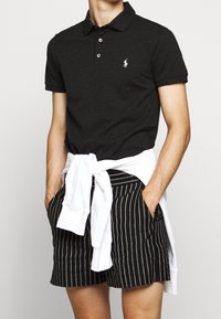 Polo noir avec un petit logo brodé, associé à un short rayé noir et blanc et un sweat-shirt blanc noué autour de la taille.