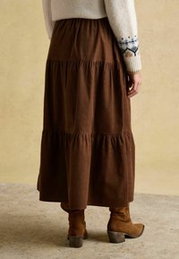 Brun maxikjol i flera lager av mjukt tyg, med en rynkad midjeband och subtil textur, kombinerad med bruna knähöga stövlar.